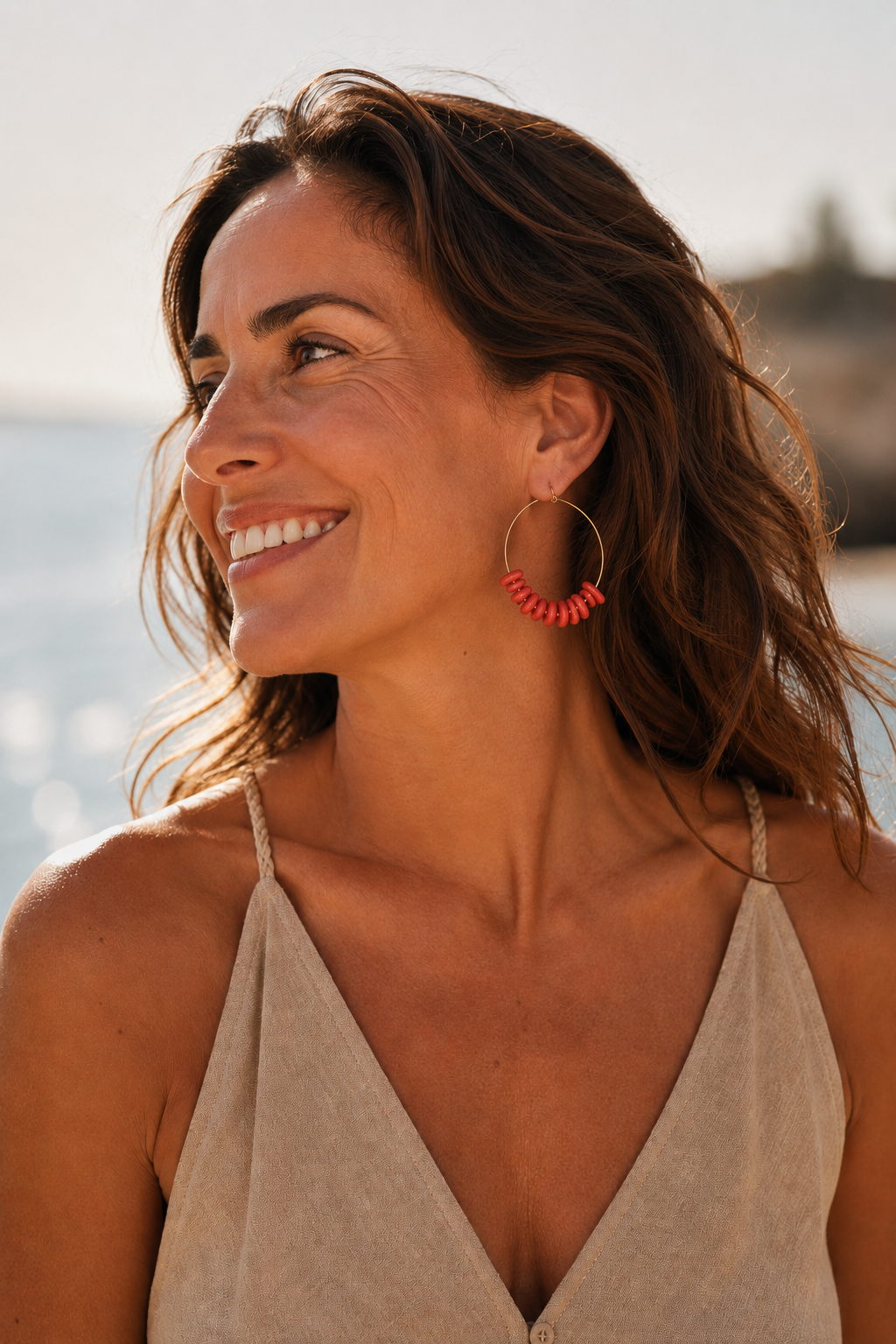 Modelo morena con vestido beige llevando los pendientes Aros Coral de Sanuke en un entorno mediterráneo junto al mar.
